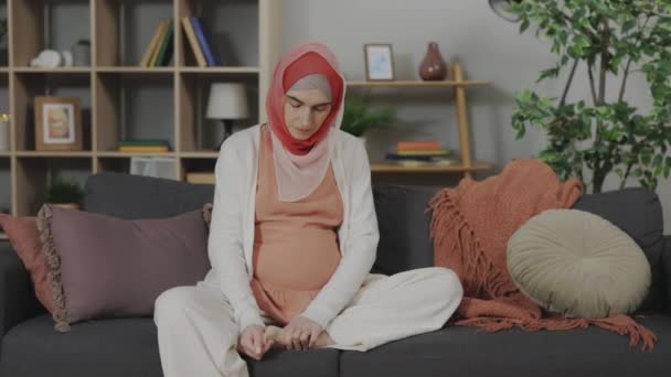 リビングルームでソファに座っている間 カジュアルな服やスカーフで妊娠中のアラビア人女性は足をマッサージ 妊娠中のリラクゼーションの概念 — ストック動画