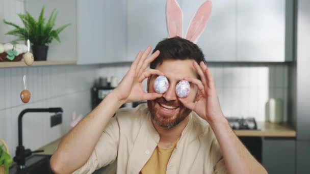 幸せハンサムな男ウサギの耳を着て 彼の目の前でカラフルなイースターエッグを保持し カメラで楽しんでいます 休日の準備 ハッピーイースター — ストック動画