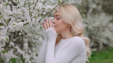 Beautiful relaxed woman enjoying nice smell of white flowers in blooming spring garden. Adorable caucasian lady running her fingers over blooming twig and looking at camera.