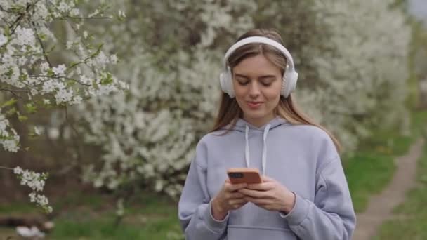 屋外を歩きながらお気に入りの歌を楽しむリラックスした若い女性のヘッドフォン 白人笑顔ブロンド手でスマートフォンを使用して無料時間を過ごす公園で満開の木々の周り — ストック動画