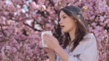 Sick caucasian woman using white napkins during runny nose outdoors. Side view of brunette standing near blooming sakura tree and suffering from seasonal allergy.