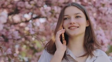 Charming young caucasian woman enjoying pleasant mobile conversation talking smartphone while standing outside near sakura tree with pink blooming. Concept of people and technology communication