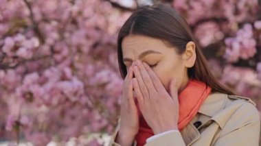 Displeased young woman rubbing eyes because of pollen allergy during spring flowering outdoors. Caucasian brunette having health problems during springtime.