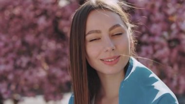 Portrait of young caucasian woman with symbolic blue and yellow ukrainian flag smiling on camera while standing outdoors. Pretty Ukrainian brunette girl posing over blooming sakura tree.
