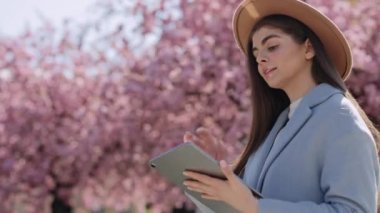 Adorable young lady with dark long hair using digital tablet while walking in the park on tje blooming sakura background. Caucasian woman wearing trendy beige hat and blue jacket