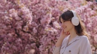Carefree young woman walking and dancing in the blossom garden while listening music in wireless headphones. Beautiful girl enjoying leisure time outdoors using modern gadget