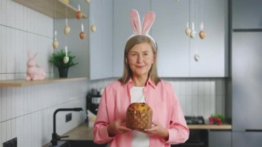 Portrait of the happy senior woman wearing bunny ears headband standing in the decorated kitchen holding the easter cake. Holiday atmosphere. Smiling mature lady preparing for easter