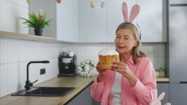 Elderly lady wearing rabbit ears sitting in the kitchen table and eating easter cake. Funny bunny senior woman holds an Easter cake in her hands. Happy female celebrating easter