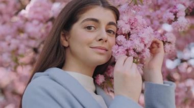 Mavi ceketli, güzel yüzünün yanında sakura ağacının dalını tutan ve kameraya bakan sevimli genç bir kadın. Dışarıda zaman geçirmek. Çiçek açan mevsimlik güzellik.