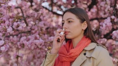 Açık havada burun spreyi kullanan, yağmurluk ve eşarp giyen koyu renk saçlı beyaz bir kadın. Sakura ağacının yanında duran hasta genç dişi bahar çiçekleriyle.