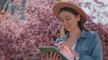Temiz hava için dijital tablet kullanan bej şapkalı ve kot gömlekli şık genç bir kadın. Gülümseyen güzel bayan baharın açtığı Sakura ağacının yanında duruyor..