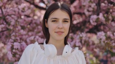 İlkbahar çiçekleri sırasında Sakura ağacının yanında poz verirken kameraya gülümseyen siyah saçlı çekici genç bir kadının portresi. Dışarıda boş vakit geçirmek için. Güzel genç yüz..