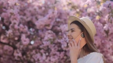 Bahçede yürüyen, arka planda çiçek açan ve cep telefonuyla konuşan beyaz kadının yan görüntüsü. Mutlu kadın esmer hasır şapka ve beyaz yaz elbisesi giyiyor..
