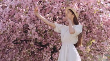 Hasır şapkalı ve beyaz yazlık elbiseli çekici bir kadın pembe çiçekli Sakura ağacının yanında selfie çekmek için modern akıllı telefon kullanıyor. İnsanlar ve teknoloji kavramı