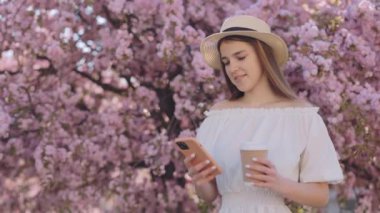 Çekici genç bir kadın elinde bir fincan kahve kâğıdıyla dışarıda dikilen ve arkasında çiçek açan sakura ağacıyla mesajlaşarak akıllı telefon kullanıyor. Gençlik yaşam tarzı için modern teknoloji