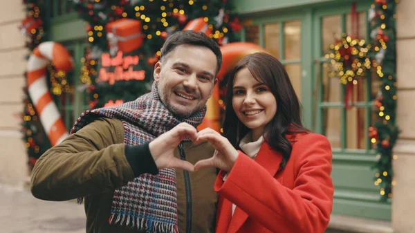 Portrait of the happy family couple making heart gesture with fingers in the decorated street, caucasian man and woman showing love hands, demonstrating sincere feelings together. Dating time