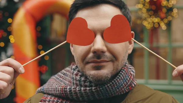 Portrait of happy smiled man playing with two paper hearts in her eyes. Pink heart, valentines day concept. Caucasian male standing outside having fun with paper hearts hiding his eyes. Close up