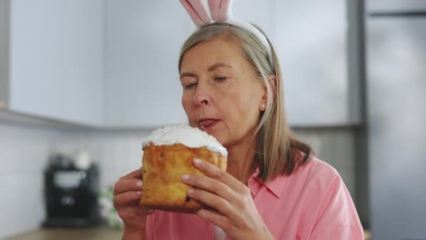 年长的女人戴着兔子耳朵坐在厨房的桌子上 享受着吃复活节蛋糕的乐趣 有趣的老女人手里拿着一个复活节蛋糕 快乐的女士庆祝复活节 — 图库视频影像