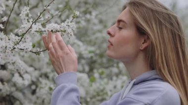 Smiling lovely Female enjoying the scent of beautiful Blossoms with closed Eyes. Positive Woman gently caressing Branch of Blooming Tree in Spring Garden. Leisure Time on Fresh Air