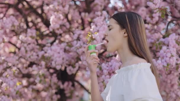 春の屋外開花の近くに喘息発作の間に吸入器を使用して長い髪を持つ白人の若い女性の側面図 季節性アレルギーの症状 — ストック動画