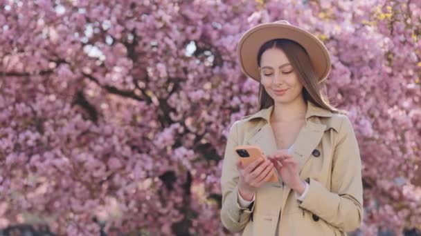 春の庭に立っている間 現代のスマートフォンを使用して帽子とトレンチコートのエレガントな若い女性 モバイル画面を見ながら笑顔魅力的な白人ブルネット 周りにピンクの桜 — ストック動画