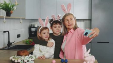 Smiling grandmother and grandchildren doing selfie with easter eggs, wearing bunny ears, have beauty and fun day together at home. Happy easter. Concept of childhood, technology, family weekend