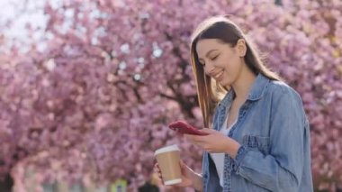 Sıradan giyinmiş çekici bir genç esmer. Akıllı telefon kullanarak lanet parkta yürüyor. Kafkasyalı Kadın Gadget Kullanıyor ve Baharın tadını çıkarıyor Sakura ağaçlarında çiçek açıyor