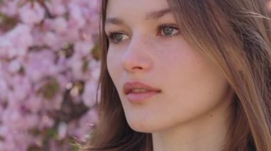 Sensual Caucasian Female Looking at Camera with Penetrated Gaze while Standing near Sakura Tree with Spring Flowering. Outdoors Portrait of Young Woman with Brown Hair. Close Up