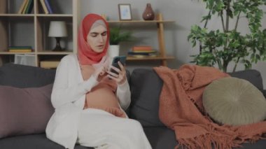 Pregnant Arabian Woman Feeling Labor Contractions While Searching Information About Babies on Cell Phone at Home. Concept of Pregnancy, Maternity and Technology