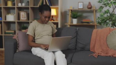 Focused Pregnant African American Woman Sitting on Sofa and Typing on Wireless Laptop. Concept of Remote Work or Remote Education During Pregnancy