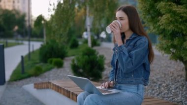 Beyaz kadın Bench 'te oturmuş, Şehir Merkezi' nde laptopun üzerinde oturmuş, kahve içiyor. Çalışan Kız Öğrenci Çalışması, Amaçlar İçin Dizüstü Bilgisayar Kullanımı. Halk ve Teknoloji Konsepti
