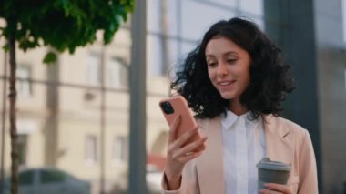 Young Businesswoman Standing Near the Business Centre Holding Coffee and Using Smartphone, Chatting with Clients While Break for Coffee. People and Technology Business Concept