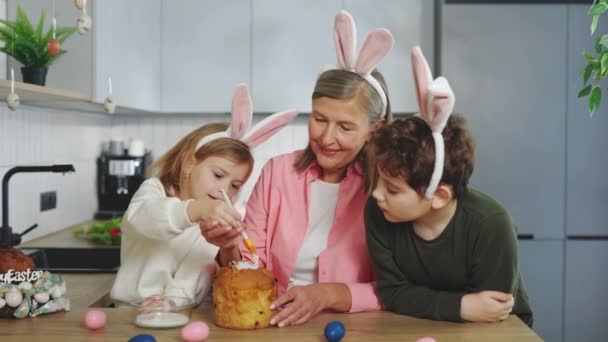 Szczęśliwa Babcia Wnukami Ubrana Królicze Uszy Dekorujące Ciasto Wielkanocne Domu — Wideo stockowe