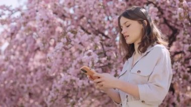 Dark-Haired Young Woman Typing Messages on Modern Smartphone while Standing Outdoors near Blooming Sakura Tree. People, Technology and Lifestyles Concept