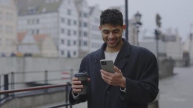 Multi-Ethnic Man Wearing a Coat, Smiling, and Walking Outside While Using Earphones and Smartphone. Winter Time. People and Technology Concept
