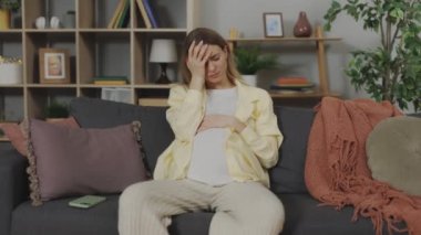 Caucasian Pregnant Woman Suffering from Headache Sitting on the Sofa at Home. Pregnancy Symptoms and Health Expectations