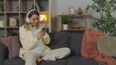 Pregnant Caucasian Woman with Smartphone in Hands Listening to Music in Wireless Headphones at Cozy Apartment. Enjoyment and Relaxation During Pregnancy Time at Home