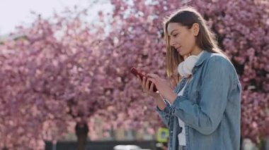 Surprised Caucasian Woman in Casual Wear Gesturing from Happiness while Getting Great News on Modern Smartphone. Joyful Brunette Walking Outdoors near Blooming Sakura Trees