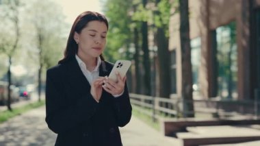 Business woman wearing formal suit standing near the business centre holding using the mobile phone, texting with clients, swiping apps. People and technology concept