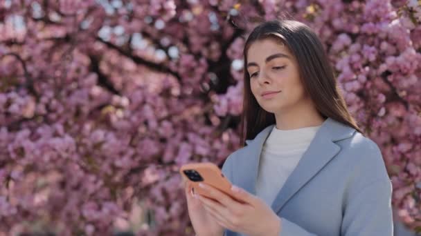 用智能手机吸引年轻的布鲁内特穿着便服站在盛开的公园里 高加索妇女移动技术 包括移动应用程序 社交媒体和讯息 — 图库视频影像