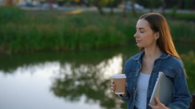 Beautiful Caucasian Female Student Walking, Holding the Laptop After Studying Drinking Coffee, Walking Near the Local Lake. Business Woman Is Going Home after Hard Work Day Enjoying Life