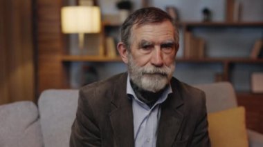 Portrait of Senior Old Gray-haired Man Sitting in the Living Room. Calm Mature Gentleman Looking at Camera at Home Interior. Elderly Senior Relaxed on the Couch. Older People Health Care