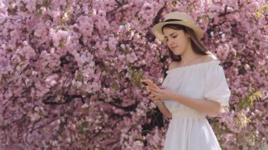 Portrait of Young Attractive Woman Standing Near Sakura Tree with Modern Smartphone in Hands and Smiling on Camera. Dark Haired Girl Wearing Straw Hat and White Summer Dress Posing Outside
