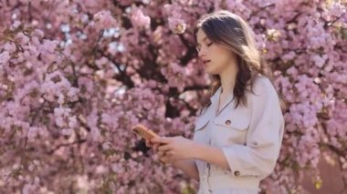 Modern Akıllı Telefonlu, Gülümseyen ve Temiz Hava 'da Kameraya Bakan Sevimli Genç Kadın. Pozitif Kafkas Esmer Pozisyon Sakura Ağacı 'nın Portresi