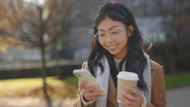 スマートフォンとテキストを使用してコーヒーカップで笑顔アジアの女の子 外で携帯電話を使っている美しい女子学生 電話で魅力的なビジネス女性 人と技術の概念 — ストック動画