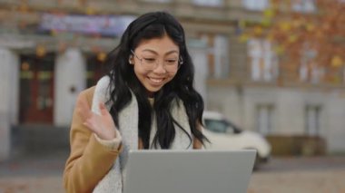 Smiled Attractive Asian Student Sitting in the Bench near University, Using Laptop, Having Distant Lecture, Waving at the Camera Using Earphones. Girl Studying Online. People and Technology Concept