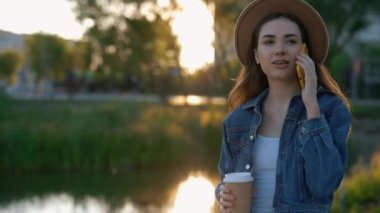 Beautiful Caucasian Woman Walking in the Park, Wearing Hat Talking on the Phone with Friends Holding Eco Cup. Attractive Lady with Braces on a Walk, Calling Someone. Female Tourist