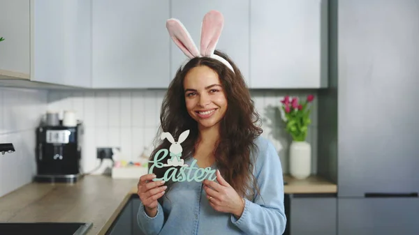 Happy Easter. Portrait of the smiling woman wearing rabbit ears headband holding the easter sign the decorated kitchen. Holiday atmosphere. Happy lady preparing for easter