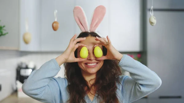 Funny brunette woman with rabbit ears on her head standing in the decorated kitchen plays with easter yellow eggs. Cute lady having fun at the camera. Preparation for the holiday.Happy Easter