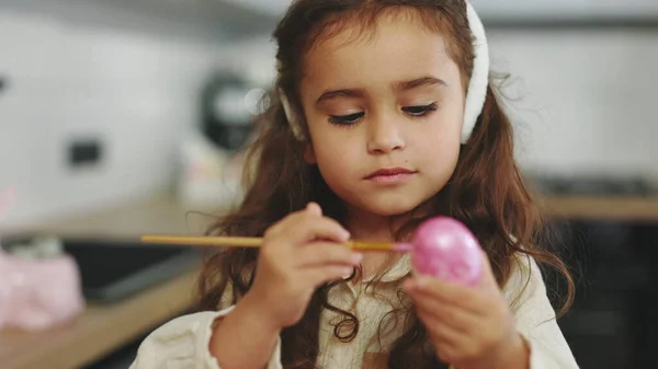 Beautiful girl painting eggs with pink paint for traditional spring dinner. Kid making easter decoration at home. Cute child wearing bunny rabbit ears on easter day preparing for holiday. Close up